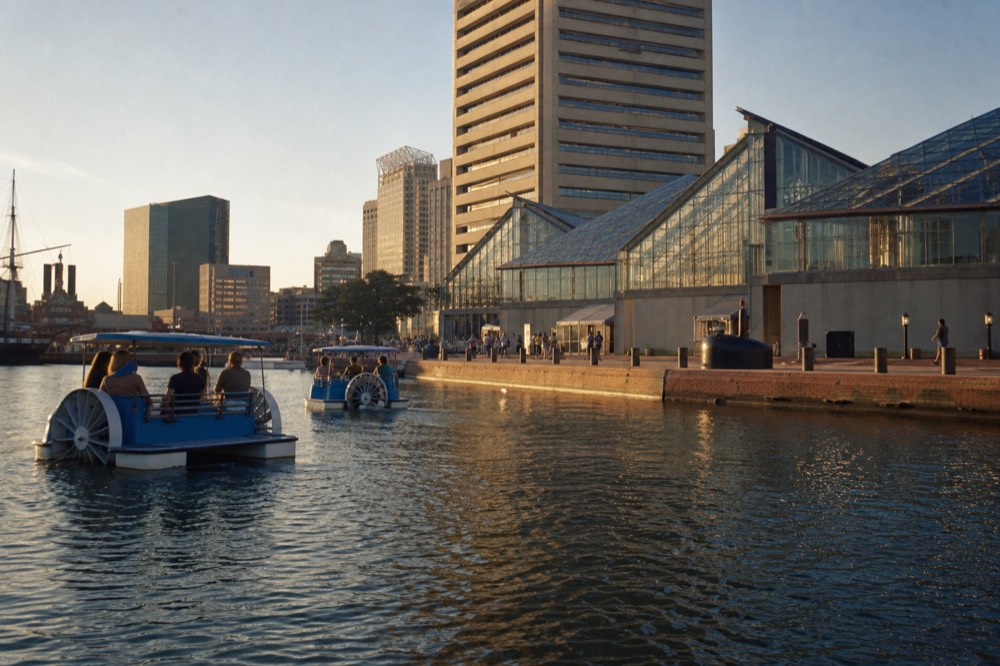 The Inner Harbor promenade seen from the water, paddleboats and glass aquarium pavilions along the brick waterfront under a bright summer sky