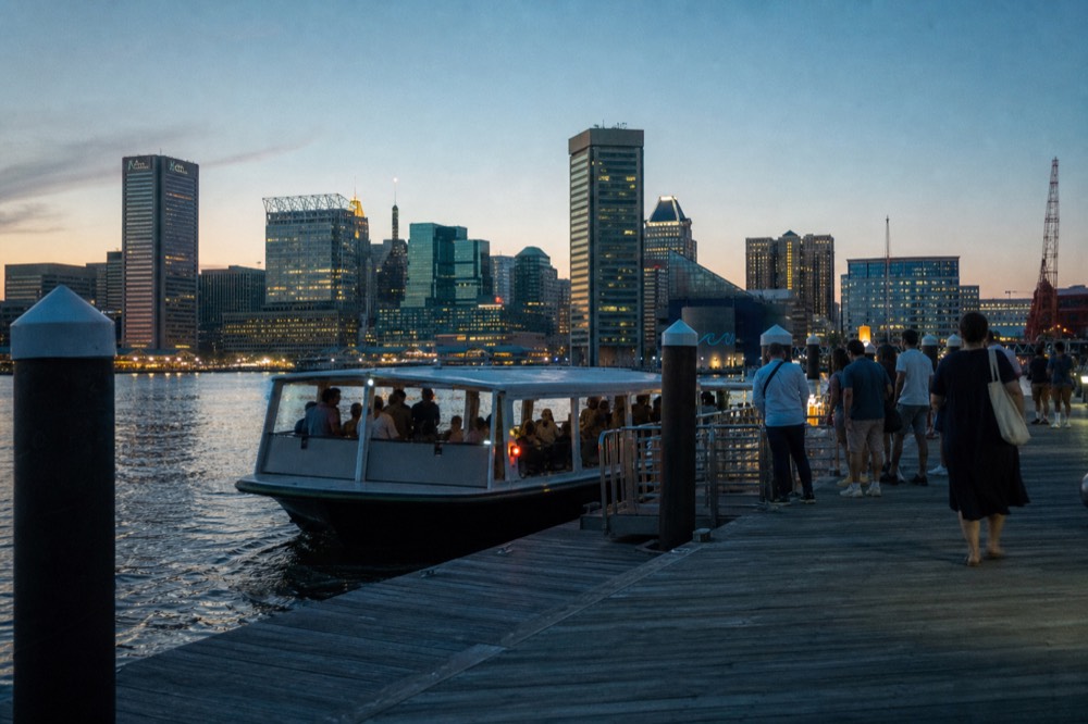 A harbor shuttle pulling up to a floating dock with a short line of riders waiting on the wooden boardwalk, late summer afternoon light