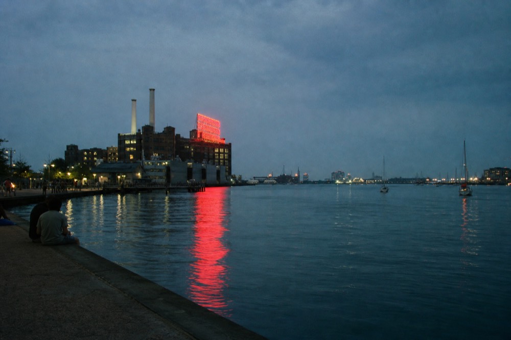 The Domino Sugars neon sign glowing red on the Locust Point waterfront at dusk, its reflection stretching across dark harbor water