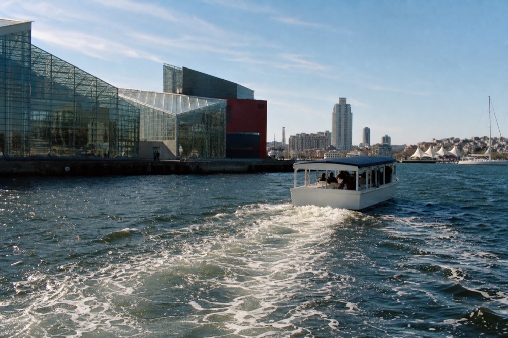 A covered narrated motor cruise boat passing the National Aquarium glass pavilions, a wide wake spreading behind it across the Inner Harbor