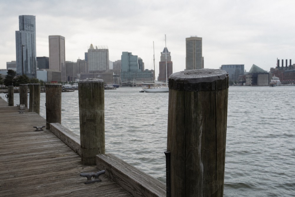 An empty timber cruise berth at the waterfront with mooring cleats and worn pilings in the foreground, the open harbor and distant skyline beyond under a pale sky