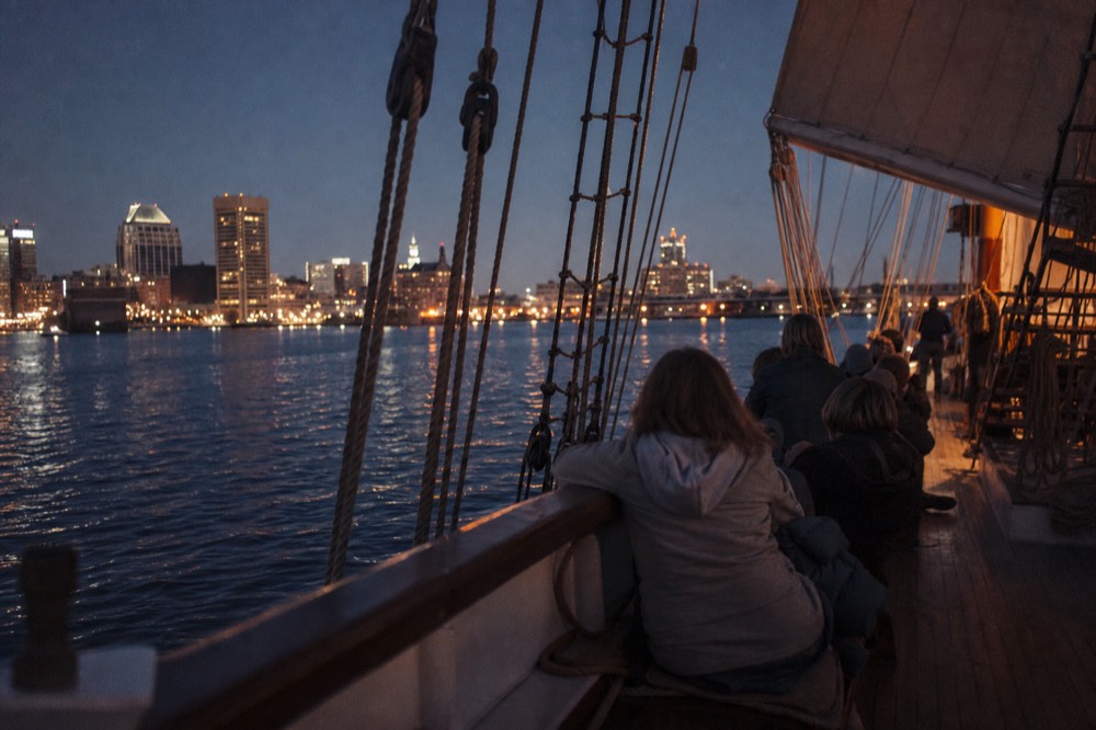 The open deck of a schooner at blue hour with a small group seated along the wooden rail, city lights glittering across the dark water behind coiled rigging