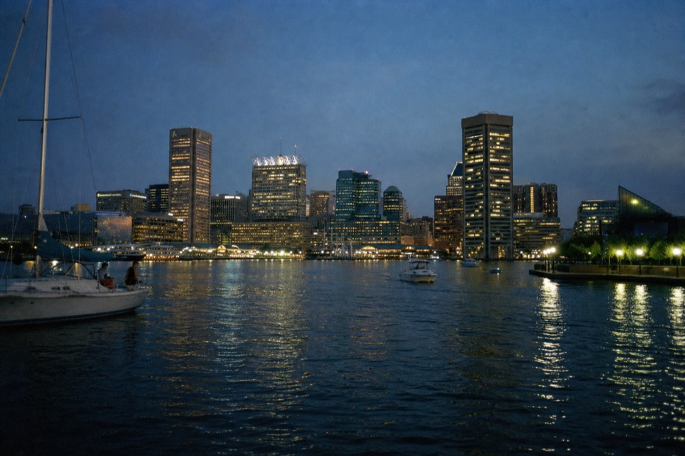 A wide view of the downtown skyline from mid-harbor at blue hour, office towers lit warm against a darkening sky, the water below scattered with the reflections of city lights