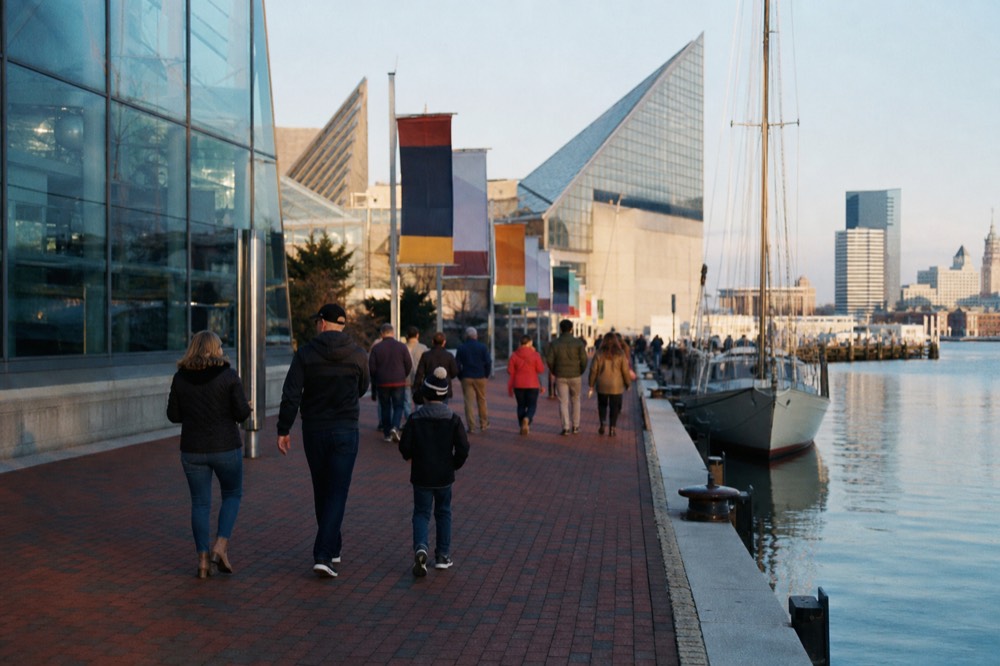 Families walking the brick promenade beside the glass pavilions of a large waterfront aquarium, colorful banners and moored boats along the edge on a clear day