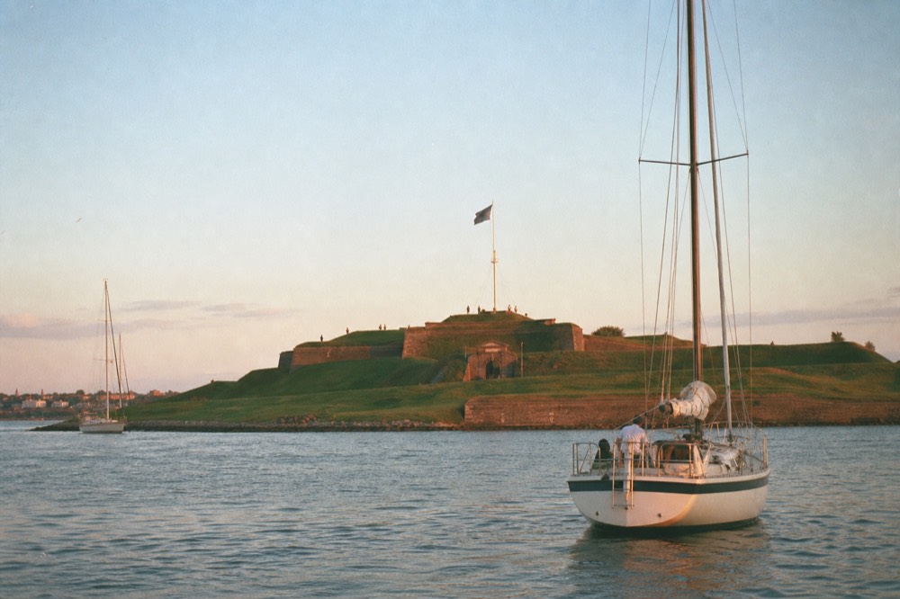 A star-shaped brick fort on a green point at the harbor mouth seen from the water, a large flag on a tall pole above the ramparts under a wide summer sky