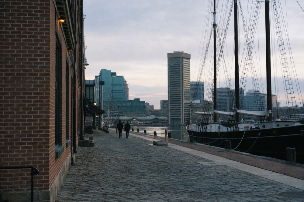 A cobblestone waterfront street of brick warehouses ending at a pier, the masts and rigging of a moored tall ship rising against a late-afternoon sky