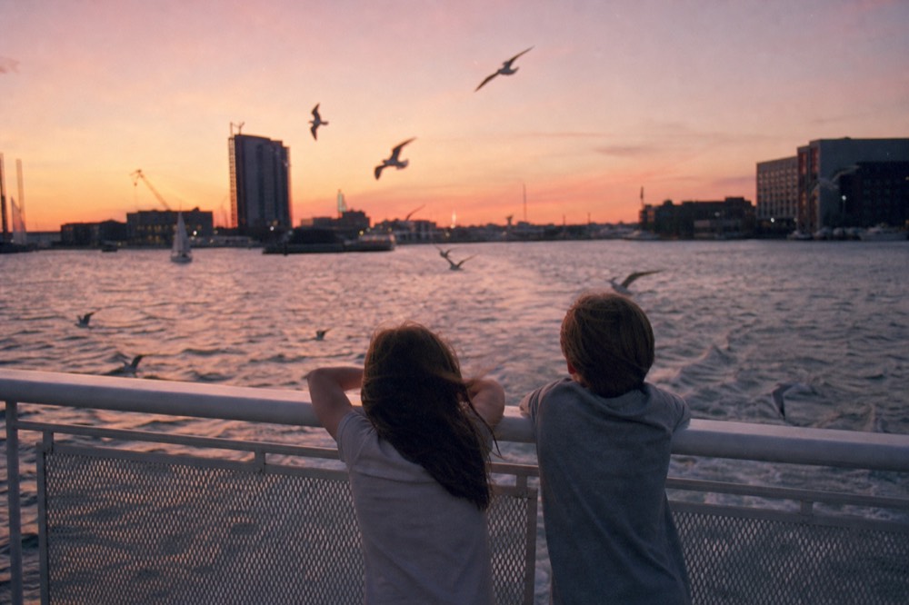 Two children leaning on the rail of an open boat deck watching the water, gulls hanging in the air above the wake, harbor buildings soft in the background