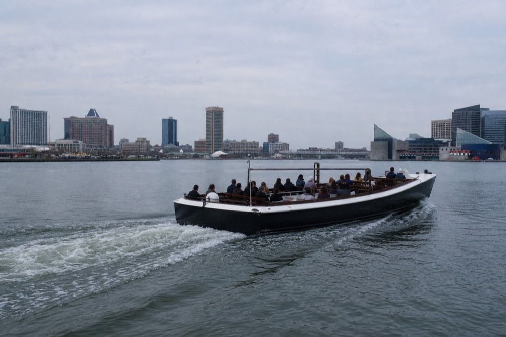 A sturdy custom-built open passenger launch with a twin-diesel wake churning behind it, crossing the Inner Harbor under a clear sky, no markings on the dark-and-white hull