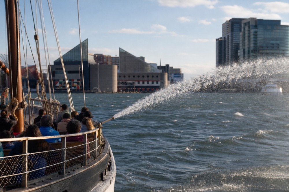 A water cannon spraying an arc of harbor spray off the side of an open excursion boat, sunlight catching the mist, families along the rail blurred in the distance