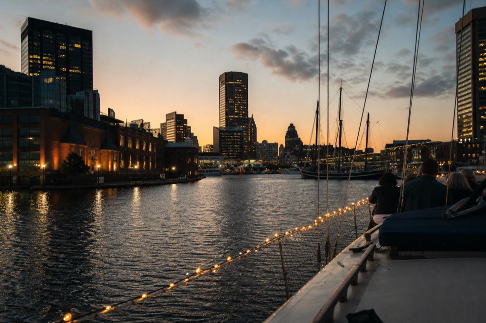 The Inner Harbor skyline at dusk seen from the deck of a cruising boat, string lights along the rail, dark water reflecting office-tower lights
