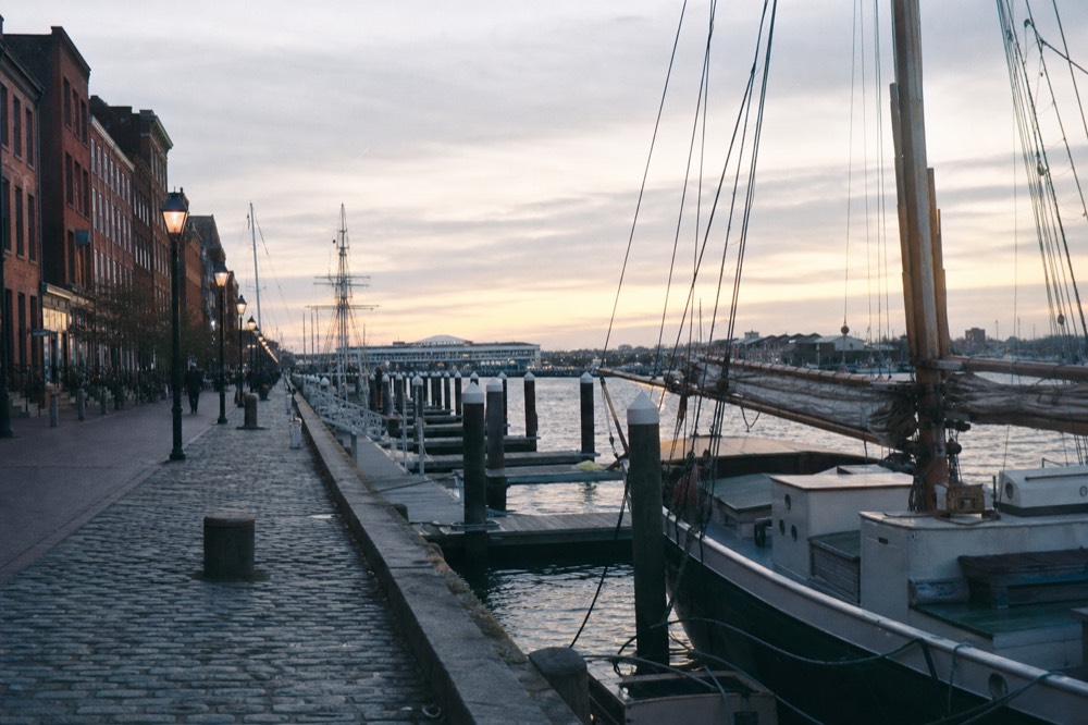 The Fells Point waterfront with floating docks and tall-ship rigging against cobblestone streets and brick rowhouses, soft evening light on the water
