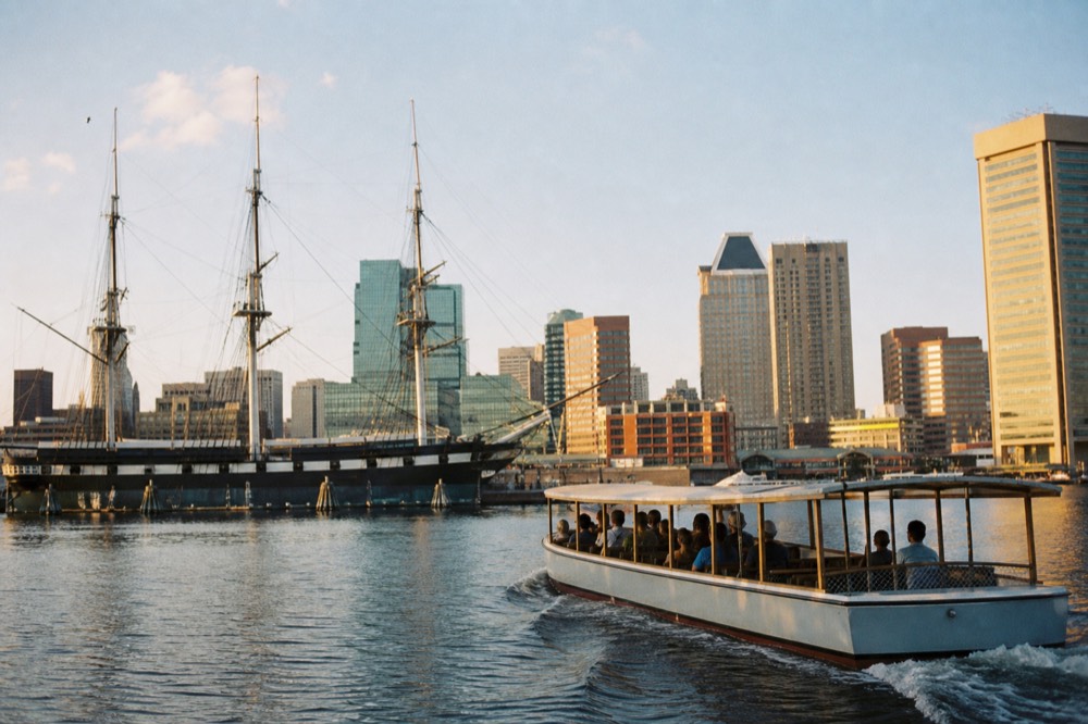 A covered narrated motor cruise boat passing the tall masts of a historic sloop-of-war in the Inner Harbor, a spreading wake across calm water under a bright sky