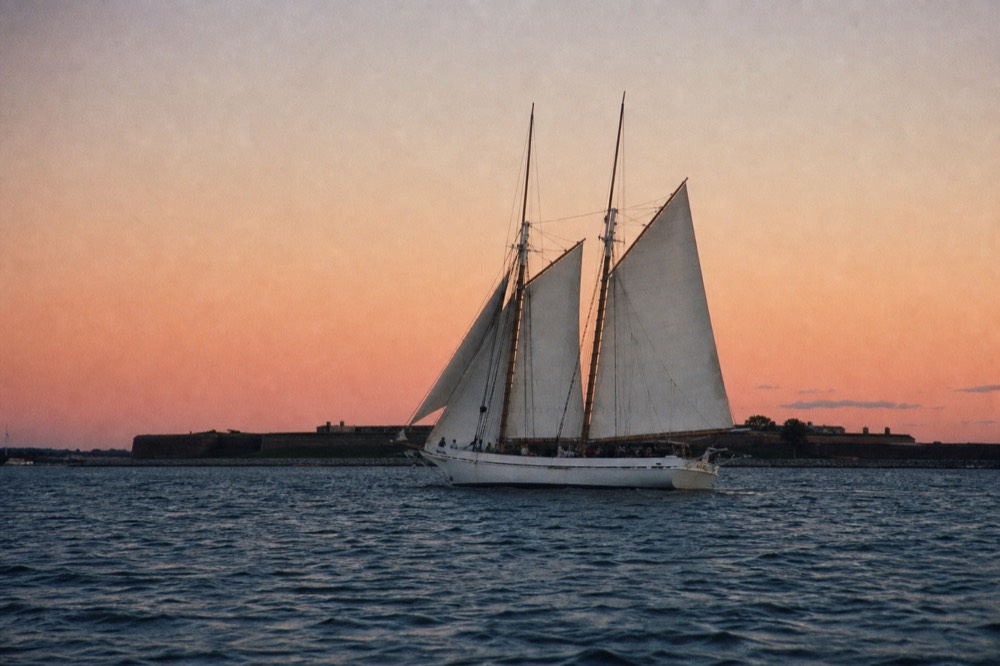 A white schooner heeling under full sail off Fort McHenry, the low star-fort ramparts on the horizon, a steady breeze filling the canvas